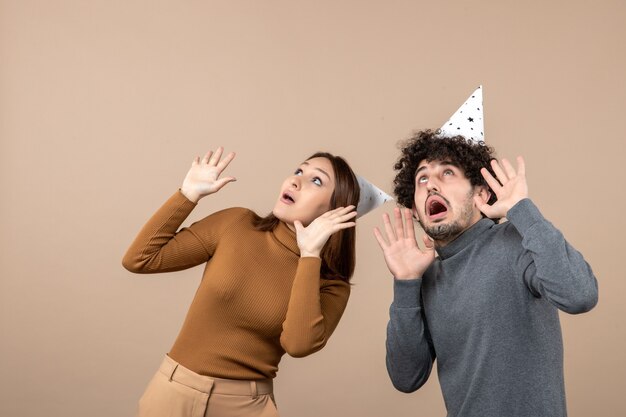 Il concetto di nuovo anno con la giovane coppia spaventata indossa il cappello del nuovo anno guardando sopra la ragazza e il ragazzo scioccati su gray