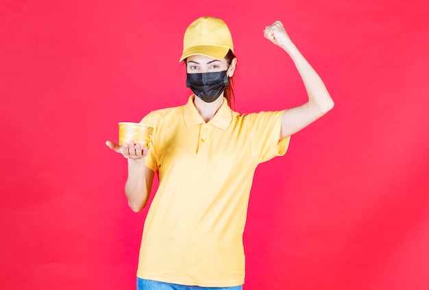 Il corriere femminile in uniforme gialla e maschera nera sta consegnando una tazza di noodles e mostrando il suo pugno