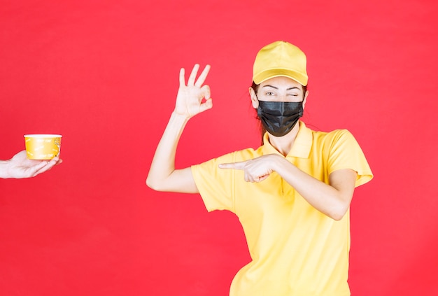 Il corriere femminile in uniforme gialla e maschera nera sta ricevendo una tazza di noodles per la consegna e mostra un segno positivo con la mano