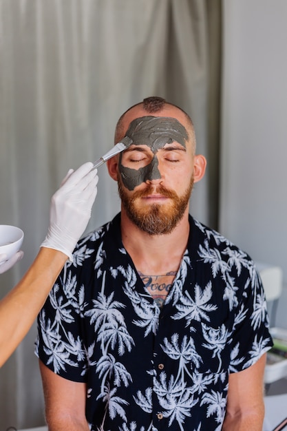 Il cosmetologo applica la maschera all'argilla con il pennello sul viso del giovane bello nella clinica di bellezza
