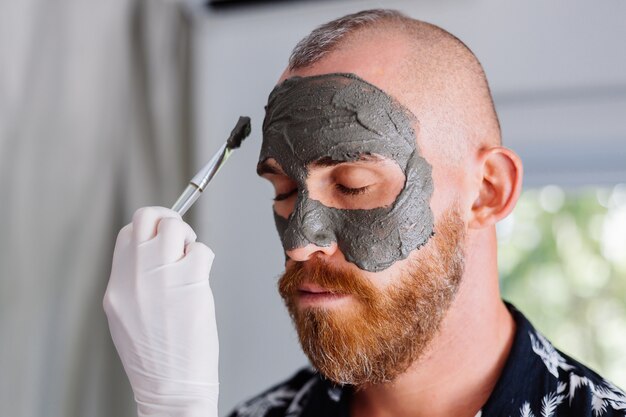 Il cosmetologo applica la maschera all'argilla con il pennello sul viso del giovane bello nella clinica di bellezza