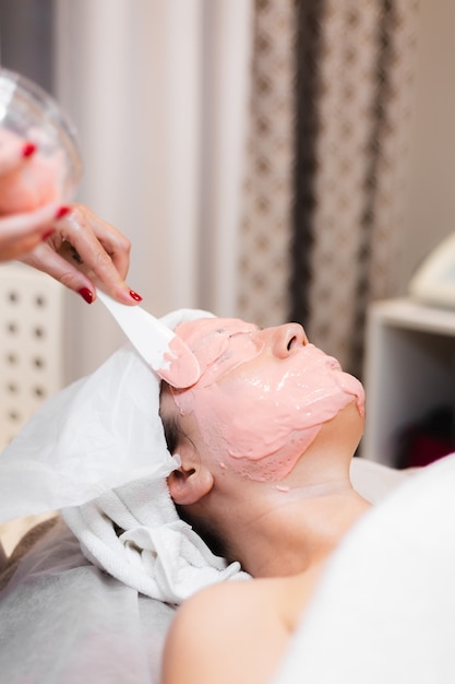 Il cosmetologo applica la maschera di alginato con la spatola sul viso della donna.