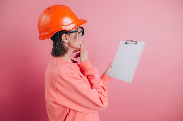 Il costruttore dell'operaio della donna abbastanza premuroso tiene il bordo bianco del segno in bianco contro fondo rosa. Casco da costruzione.