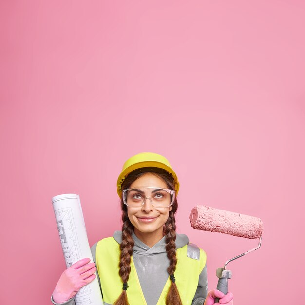 Il costruttore o l'architetto donna soddisfatto tiene la testa al sicuro nel casco indossa occhiali protettivi tiene il rullo di vernice e fornisce il progetto il miglior servizio di sempre utilizza lo strumento per la riparazione pronto per le sfide