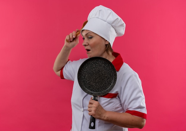 Il cuoco femminile di mezza età confuso in padella della tenuta dell'uniforme del cuoco unico ha messo il cucchiaio nella sua fronte sulla parete rosa isolata