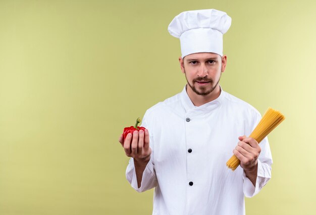 Il cuoco unico maschio professionista soddisfatto cucina in uniforme bianca e cappello del cuoco che tiene il peperone fresco e della pasta degli spaghetti crudi che sta sopra il fondo verde