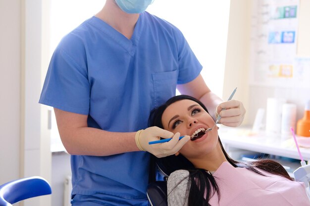 Il dentista in uniforme blu e guanti bianchi si prende cura di una donna caucasica bruna sexy, sta dietro di lei, guarda nella sua bocca con un piccolo specchio.