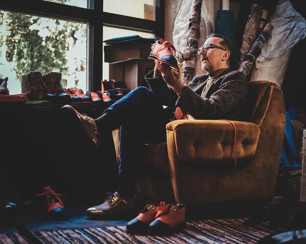 Il designer di scarpe maturo è seduto sulla poltrona del suo laboratorio e tiene in mano una scarpa della sua ultima collezione.