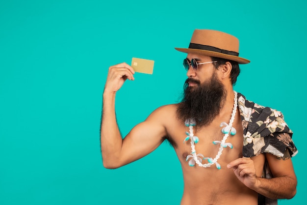 Il di un uomo felice barba lunga che indossa un cappello, indossa una camicia a righe, in possesso di una carta di credito dorata su un blu.