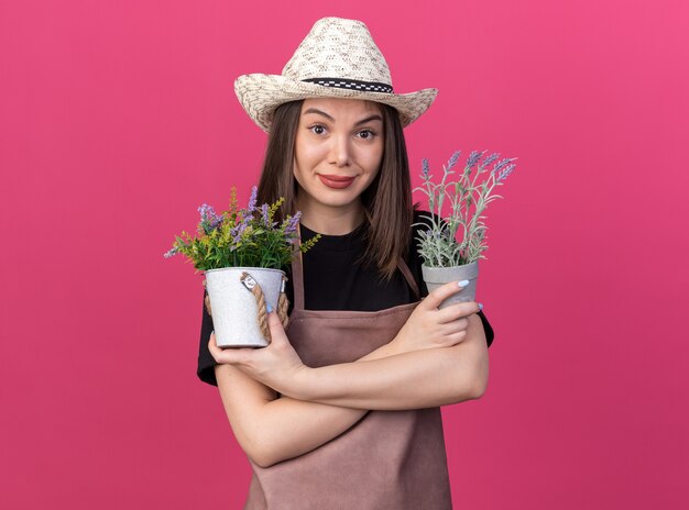 Il giardiniere femminile abbastanza caucasico piacevole che indossa il cappello da giardinaggio sta con le braccia incrociate che tengono i vasi di fiori isolati sulla parete rosa con lo spazio della copia