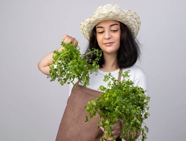 Il giardiniere femminile giovane brunetta soddisfatto in cappello da giardinaggio d'uso uniforme tiene ed esamina il coriandolo isolato sulla parete bianca