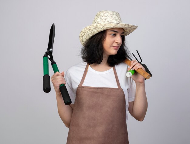 Il giardiniere femminile giovane brunetta soddisfatto in cappello da giardinaggio d'uso uniforme tiene gli attrezzi da giardinaggio che esaminano lato isolato sulla parete bianca