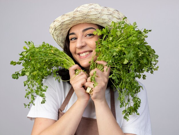 Il giardiniere femminile giovane brunetta soddisfatto in cappello da giardinaggio da portare uniforme tiene il coriandolo isolato sulla parete bianca con lo spazio della copia