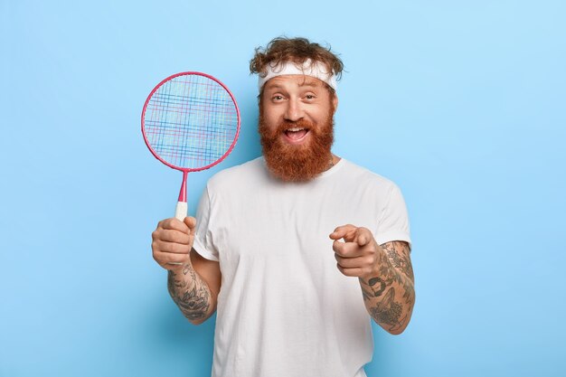 Il giocatore di tennis dai capelli rossi amichevole allegro tiene la racchetta mentre posa contro il muro blu