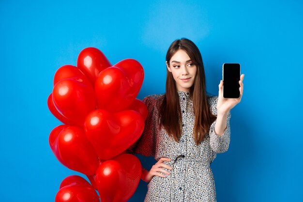 Il giorno di San Valentino bella donna sorridente in abito che mostra lo schermo vuoto dello smartphone in piedi vicino a un romantico ...