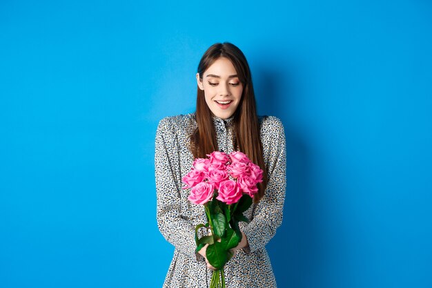 Il giorno di San Valentino concetto. Felice donna attraente riceve fiori a sorpresa, guardando grato al bouquet di rose rosa, in piedi su sfondo blu.