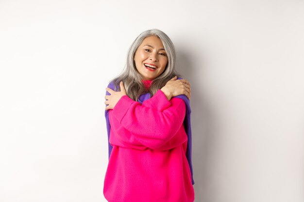 Il giorno di San Valentino e il concetto di vacanze. Bella donna asiatica anziana in maglione rosa che si abbraccia con gli occhi chiusi, sorridente, in piedi su sfondo bianco
