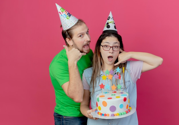 Il giovane allegro che indossa i gesti del cappello del partito mi chiama segno in piedi con la ragazza sorridente che indossa il cappello del partito e che tiene la torta di compleanno che gesticola mi chiama segno isolato sul muro rosa