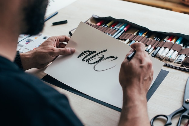 Il giovane artista illustratore hipster in maglietta semplice nera crea scritte a mano autentiche e uniche nel suo luminoso studio industriale