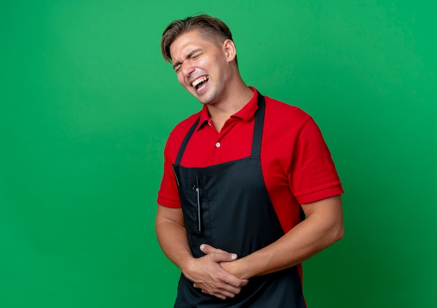 Il giovane barbiere maschio biondo allegro in uniforme mette le mani sulla pancia isolata su spazio verde con lo spazio della copia