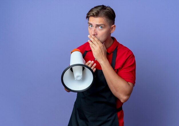 Il giovane barbiere maschio biondo ansioso in uniforme mette la mano sulla bocca che tiene altoparlante isolato sullo spazio viola con lo spazio della copia