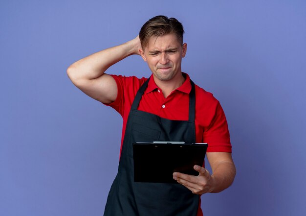 Il giovane barbiere maschio biondo ansioso in uniforme tiene la testa dietro guardando appunti isolato sullo spazio viola con lo spazio della copia