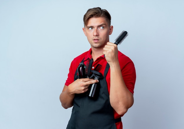 il giovane barbiere maschio biondo confuso in uniforme tiene gli strumenti del barbiere che esaminano lato