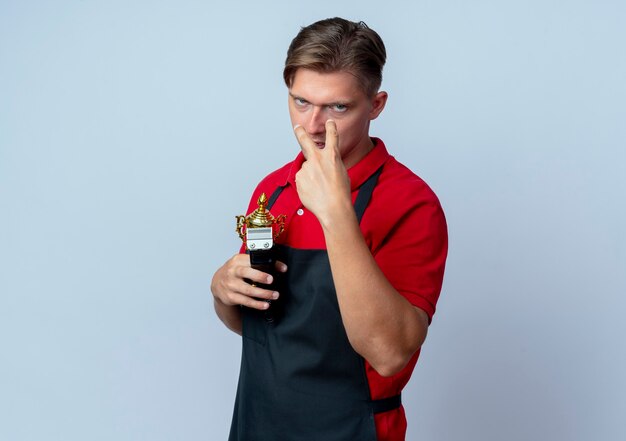 Il giovane barbiere maschio biondo furioso in uniforme tiene il tagliatore di capelli e la tazza del vincitore che indica agli occhi isolati su fondo bianco con lo spazio della copia