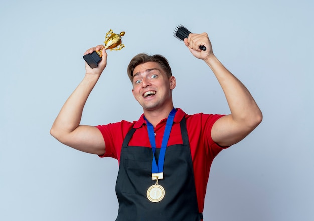 Il giovane barbiere maschio biondo gioioso in uniforme con la medaglia d'oro tiene la tazza del vincitore e il pettine isolato su uno spazio bianco con lo spazio della copia