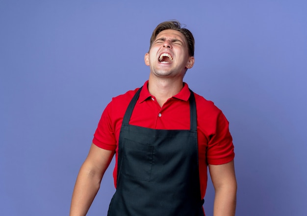 Il giovane barbiere maschio biondo infastidito in uniforme grida con gli occhi chiusi isolati sullo spazio viola con lo spazio della copia