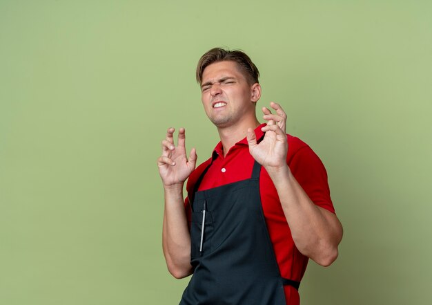 Il giovane barbiere maschio biondo infastidito in uniforme stringe le mani isolate sullo spazio verde oliva con lo spazio della copia