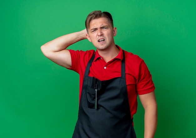 Il giovane barbiere maschio biondo infastidito in uniforme tiene la testa dietro isolato su spazio verde con lo spazio della copia