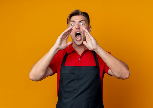 Il giovane barbiere maschio biondo infastidito in uniforme tiene le mani vicino alla bocca fingendo di chiamare qualcuno isolato sullo spazio arancione con lo spazio della copia