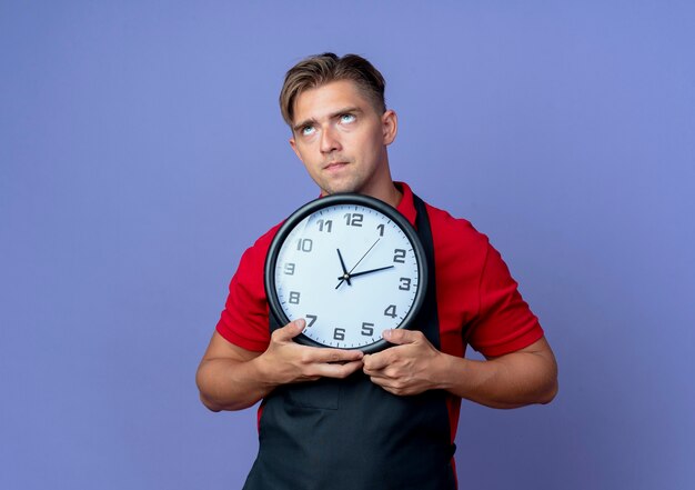 Il giovane barbiere maschio biondo premuroso in uniforme tiene l'orologio che osserva in su isolato sullo spazio viola con lo spazio della copia