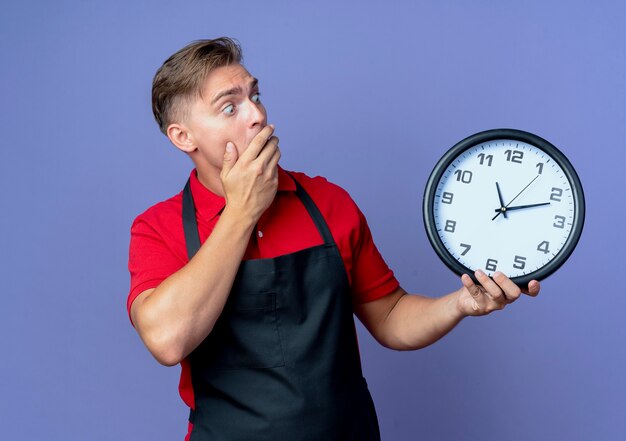 Il giovane barbiere maschio biondo scioccato in uniforme tiene ed esamina l'orologio isolato sullo spazio viola con lo spazio della copia