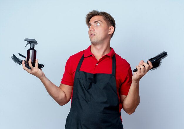 Il giovane barbiere maschio biondo scioccato in uniforme tiene gli strumenti del barbiere che esaminano lato isolato su spazio bianco con lo spazio della copia