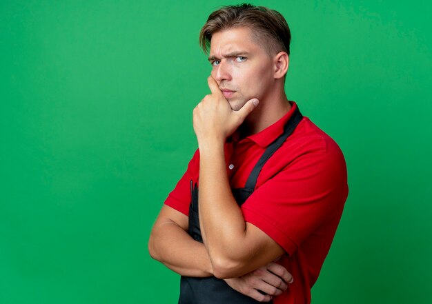 Il giovane barbiere maschio biondo serio in uniforme mette la mano sul mento isolato su spazio verde con lo spazio della copia