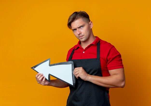 Il giovane barbiere maschio biondo serio in uniforme tiene il segno di direzione isolato sullo spazio arancio con lo spazio della copia