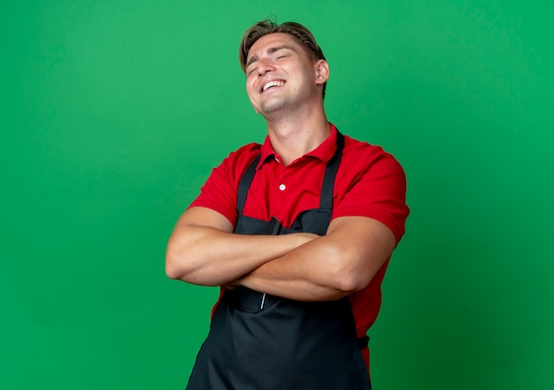 Il giovane barbiere maschio biondo sorridente in uniforme sta con le braccia incrociate isolato su spazio verde con lo spazio della copia