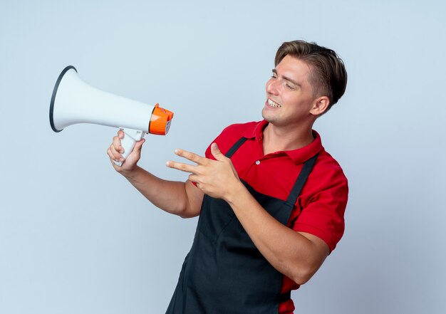 Il giovane barbiere maschio biondo sorridente in uniforme tiene l'altoparlante che esamina il lato isolato su fondo bianco con lo spazio della copia