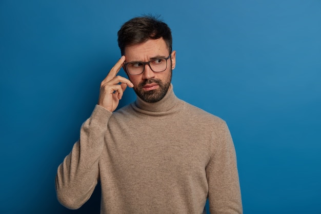 Il giovane barbuto pensieroso tiene il dito sulla tempia, ricorda le informazioni necessarie in mente, indossa occhiali trasparenti e un maglione casual