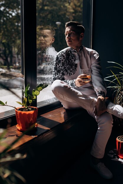 Il giovane bello in vestito bianco con ricamo si siede su un davanzale prima della finestra luminosa