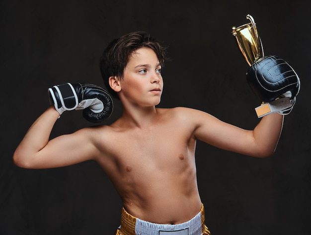 Il giovane campione di pugili senza maglietta che indossa guanti tiene una coppa del vincitore che mostra i muscoli. Isolato su uno sfondo scuro.