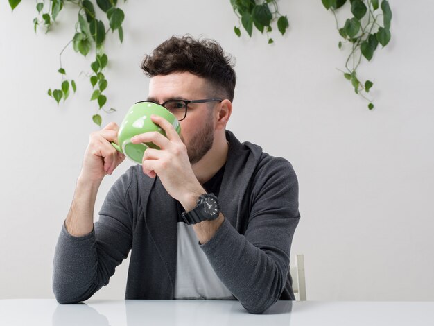 il giovane che si siede in occhiali guarda la giacca grigia che beve il suo succo dalla tazza verde insieme alla pianta su bianco