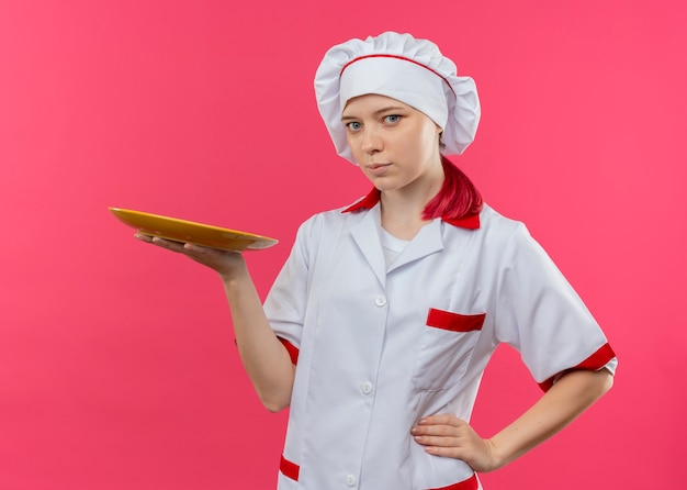 Il giovane chef femmina bionda felice in uniforme da chef tiene il piatto e mette la mano sulla vita isolata sulla parete rosa