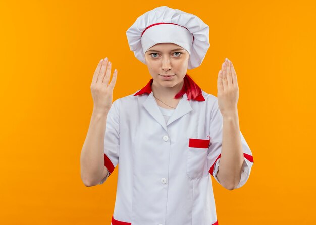 Il giovane chef femmina bionda felice in uniforme da chef tiene le mani in alto isolato sulla parete arancione