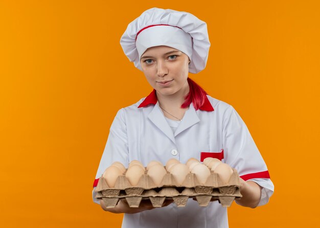Il giovane chef femmina bionda felice in uniforme da chef tiene un lotto di uova isolate sulla parete arancione