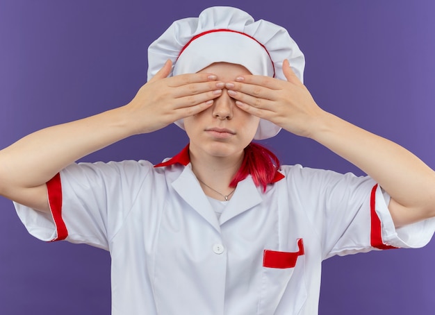 Il giovane chef femmina bionda fiducioso in uniforme da chef chiude gli occhi con le mani isolate sulla parete viola