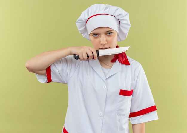 Il giovane chef femmina bionda fiducioso in uniforme da chef tiene il coltello e sembra isolato sulla parete verde
