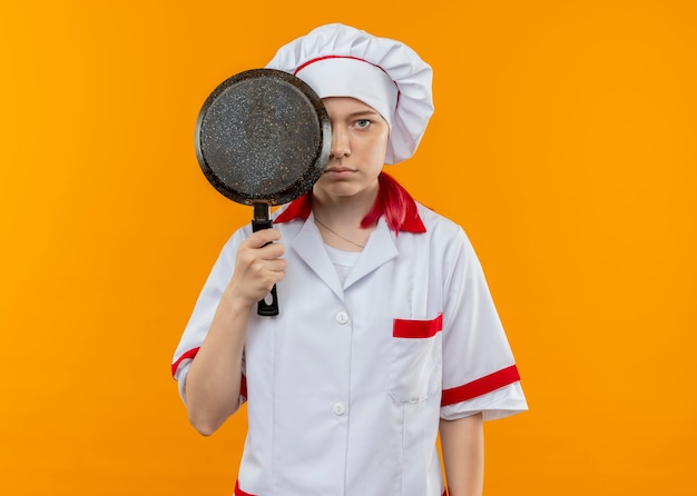 Il giovane chef femminile biondo fiducioso in uniforme del cuoco unico chiude l'occhio con la padella isolata sulla parete arancione